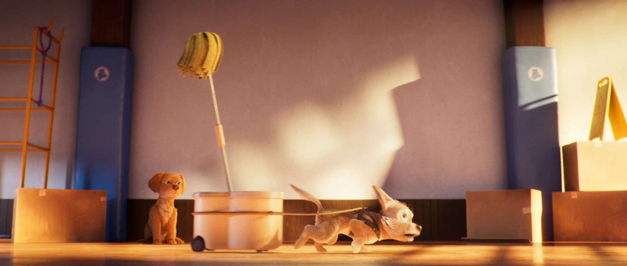 A puppy in a harness runs pulling a mop bucket while a golden puppy sits and watches.